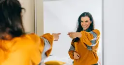 Uśmiechnięta kobieta w musztardowej bluzie wskazuje palcem na swoje odbicie w lustrze, emanując charyzmą.