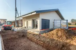 Budowa domu: wykop pod fundamenty, koparka i częściowo wykończony budynek.