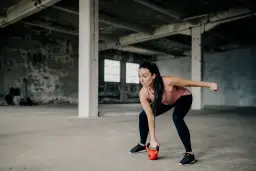 Kobieta w koszulce treningowej wykonuje ćwiczenie z kettlebell w opuszczonej hali.