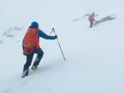 Dwóch turystów w kaskach i z plecakami wspina się po ośnieżonym zboczu, używając kijków i raków do chodzenia po górach.
