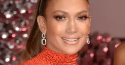 Zbliżenie na twarz Jennifer Lopez w błyszczącej, pomarańczowej sukience, z biżuterią.