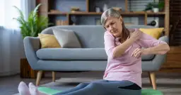 Starsza kobieta siedzi na macie do ćwiczeń, trzymając się za obolałe ramię.