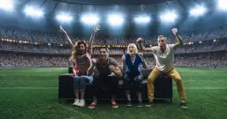 Cztery osoby siedzące na kanapie na boisku piłkarskim, kibicujące na tle stadionu.