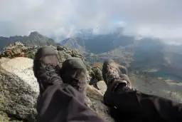 Stopy w butach trekkingowych na tle jesiennych Tatr. Październikowe słońce przebija się przez chmury, oświetlając skaliste szczyty i turkusowe jezioro.