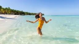 Szczęśliwa kobieta w żółtym stroju kąpielowym biegnie przez turkusową wodę na plaży z palmami w tle.