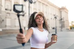 Uśmiechnięta content reviewer z kawą w ręku nagrywa vlog na ulicy przed okazałym budynkiem.