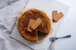 Pyszny sernik lotus z polewą karmelową i pokruszonymi ciasteczkami. Obok leżą dwa ciasteczka lotus i widelec.