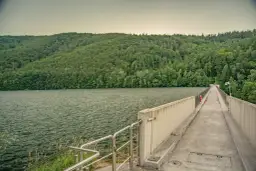 Zapora wodna z betonowym chodnikiem i balustradą, otoczona zielonymi wzgórzami i jeziorem.