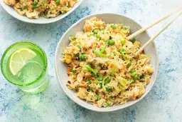 Pyszne krewetki z ryżem, groszkiem i marchewką, podane z pałeczkami. Obok szklanka orzeźwiającej wody z cytryną.