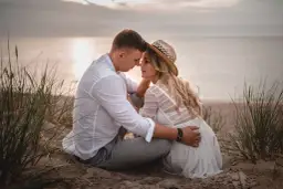 Romantyczna sesja we dwoje na plaży. Para siedzi blisko siebie, otoczona trawami, z widokiem na spokojne morze o zachodzie słońca.