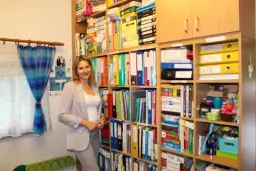 Kobieta w jasnej marynarce stoi w nowoczesnym gabinecie logopedycznym, otoczona regałami pełnymi materiałów terapeutycznych.
