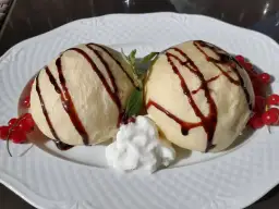 Dwa kluski na parze polane czekoladą, z bitą śmietaną i czerwonymi porzeczkami.