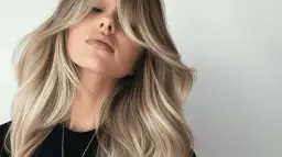 Blond włosy z grzywką na boki, ułożone prostownicą. Delikatne fale i subtelny makijaż podkreślają urodę.