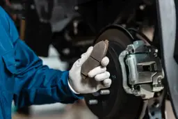Mechanik w niebieskim kombinezonie trzyma zużyty klocek hamulcowy, zastanawiając się, ile kosztuje wymiana zacisku hamulcowego.