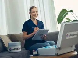 Kobieta w kitlu siedzi na kanapie, trzymając dokumenty i długopis. Obok niej stoi walizka z materiałami medycznymi.