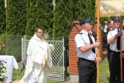 Ksiądz w białej szacie niesie monstrancję, obok stoją mężczyźni w mundurach.