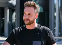 Aleksander Sikora w czarnej koszulce z plecakiem, na tle budynku.