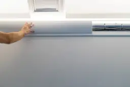 Ręka przytrzymuje białą listwę, która ukrywa kable. Pokazano, jak schować kabel w styropianie, tworząc estetyczne wykończenie.