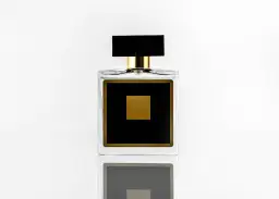 Elegancka szklana butelka perfum z czarną nakrętką i złotym kwadratem na czarnym tle.