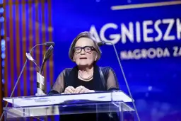 Agnieszka Holland w okularach za mównicą z mikrofonem i statuetką.