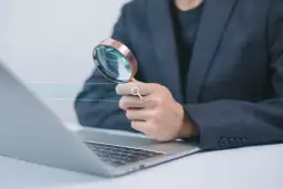 Osoba w garniturze trzyma lupę nad polem wyszukiwania na tle laptopa.
