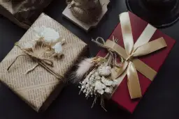 Elegancko zapakowane prezenty na urodziny: czerwone pudełko ze złotą wstążką i bukiecikiem suszonych kwiatów oraz prezent owinięty w papier z tekstem, ozdobiony sznurkiem i białymi kwiatami.