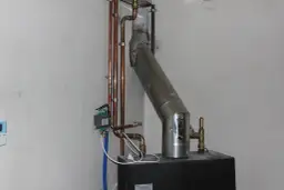 Szara masa uszczelniająca łączy rurę pieca z kominem. Widać też miedziane rurki instalacji grzewczej.