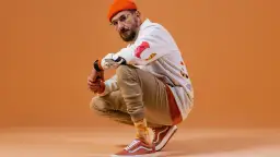 DJ BRK robię co mogę, w pomarańczowej czapce i okularach, kuca na pomarańczowym tle.