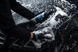 Ręce w czarnych rękawiczkach myją czarne auto pianą, trzymając butelkę szamponu.