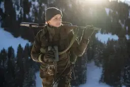 Kobieta w zimowym krajobrazie z karabinem na ramieniu i lornetką w dłoni. Czeka na to, co ustrzelił myśliwy.