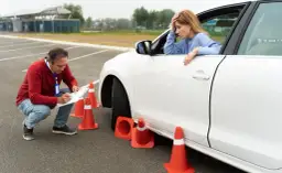 Zestresowana kursantka podczas **testów opiekunka dziecięca** na placu manewrowym. Instruktor notuje jej postępy.