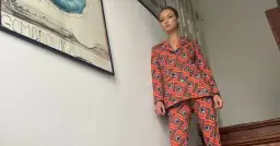 Kobieta w kolorowym, wzorzystym garniturze, przypominającym styl, jak się ubierano w latach 70. Obok plakat teatralny.