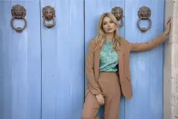 Kobieta w beżowym garniturze i wzorzystej bluzce stoi przed niebieskimi drzwiami z ozdobnymi uchwytami.