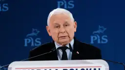 Jarosław Kaczyński w garniturze przemawia za mównicą na tle niebieskiego baneru z logo PiS.