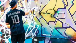 Mann mit Hut und Trikot sprüht mit Spraydose auf bunte Graffiti-Wand.