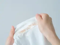 Dłonie trzymają białą bluzę z plamami podkładu. Dowiedz się, jak zmyć podkład z bluzy.