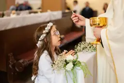 Dziewczynka w wianku na głowie klęczy podczas komunii, przyjmując hostię od księdza.