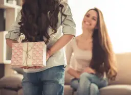 Dziecko ukrywa prezent za plecami przed uśmiechniętą kobietą.