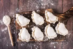 Surowe pierogi na desce, gotowe do przechowywania w lodówce. Obok łyżka z mąką.