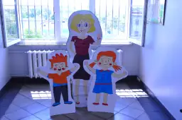 Kiedy śmieje się dziecko, świat staje się lepszy." - Korczak. Postacie z papieru: pani i dwoje dzieci, stoją przy oknie.