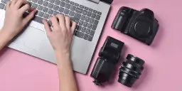 Ręce piszą na laptopie, obok aparat, lampa błyskowa i obiektyw. Szukasz, gdzie sprzedać aparat fotograficzny?