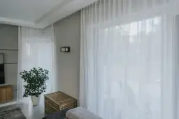 Jasne, białe zasłony firanowe zwisają z ukrytego karnisza na przelotki, tworząc delikatną atmosferę.