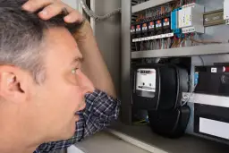 Mężczyzna zmartwiony patrzy na licznik energii elektrycznej w skrzynce z bezpiecznikami.