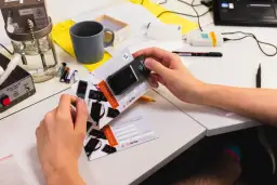 Ręce trzymają certyfikat kalibracji alkomatu. Na stole leżą baterie, kubek i sprzęt laboratoryjny. Dowiedz się, ile kosztuje kalibracja alkomatu.