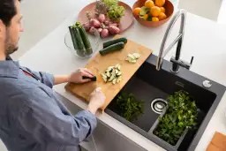 Mężczyzna kroi cukinię na desce obok czarnego zlewu z kranem i natką pietruszki.