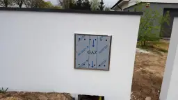 Metalowa skrzynka gazowa z napisem "GAZ" zamontowana na białej ścianie, obok niej rośnie młode drzewko.