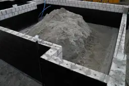 Duża hałda piasku czeka, by zasypać fundamenty z zewnątrz. Ściany fundamentów pokryte czarną izolacją.