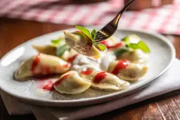 Jak gotować pierogi mrożone? Na talerzu pyszne pierogi z polewą truskawkową i miętą, jeden pieróg na widelcu.