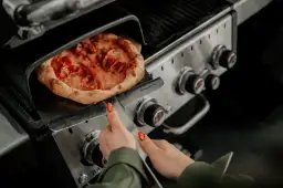 Pizza na grillu, jak piec bez kamienia. Ręka z paznokciem w kształcie pizzy zbliża się do gorącego dania.