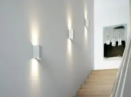 Nowoczesne oświetlenie nocne korytarza tworzy nastrojową atmosferę. Trzy białe kinkiety rzucają snopy światła na szarą ścianę, oświetlając schody i abstrakcyjny obraz.